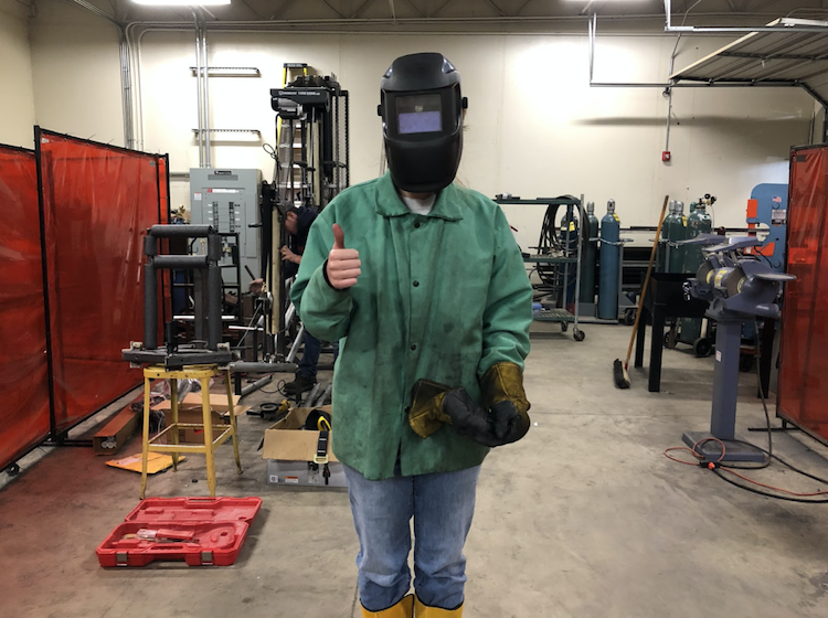 Decked out in full welding gear, Miranda Almen gives a thumbs-up in the manufacturing lab where she's learning how to MIG weld!